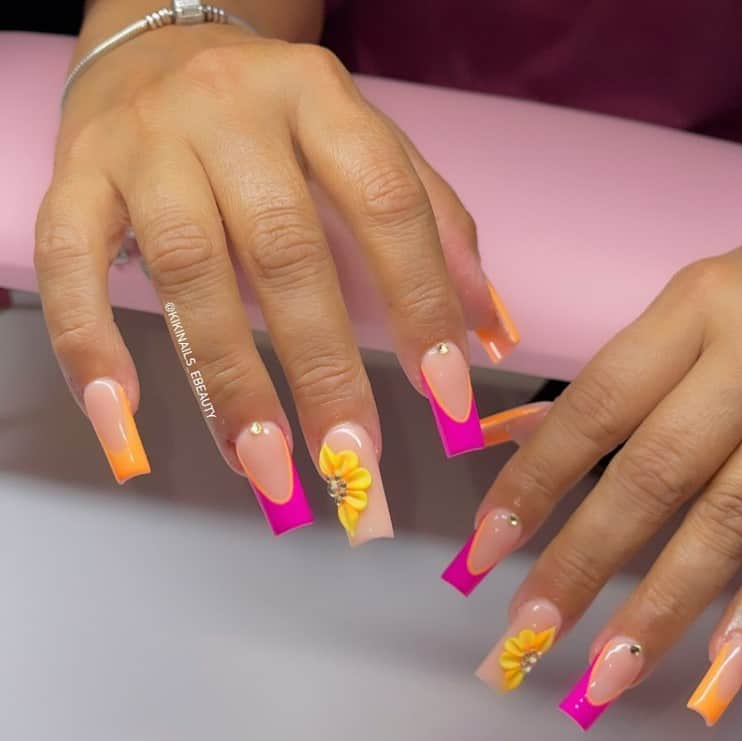 A woman's hands with peach nail polish base that has orange tips and hot pink tips and 3D yellow flowers