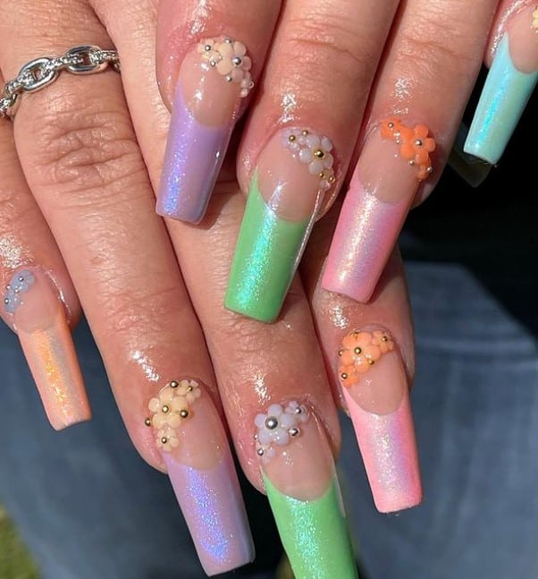 A closeup of a woman's hands with a nude nail polish base that has thicker French tips in very pale pastel colors, shimmery polish and 3D pastel flowers