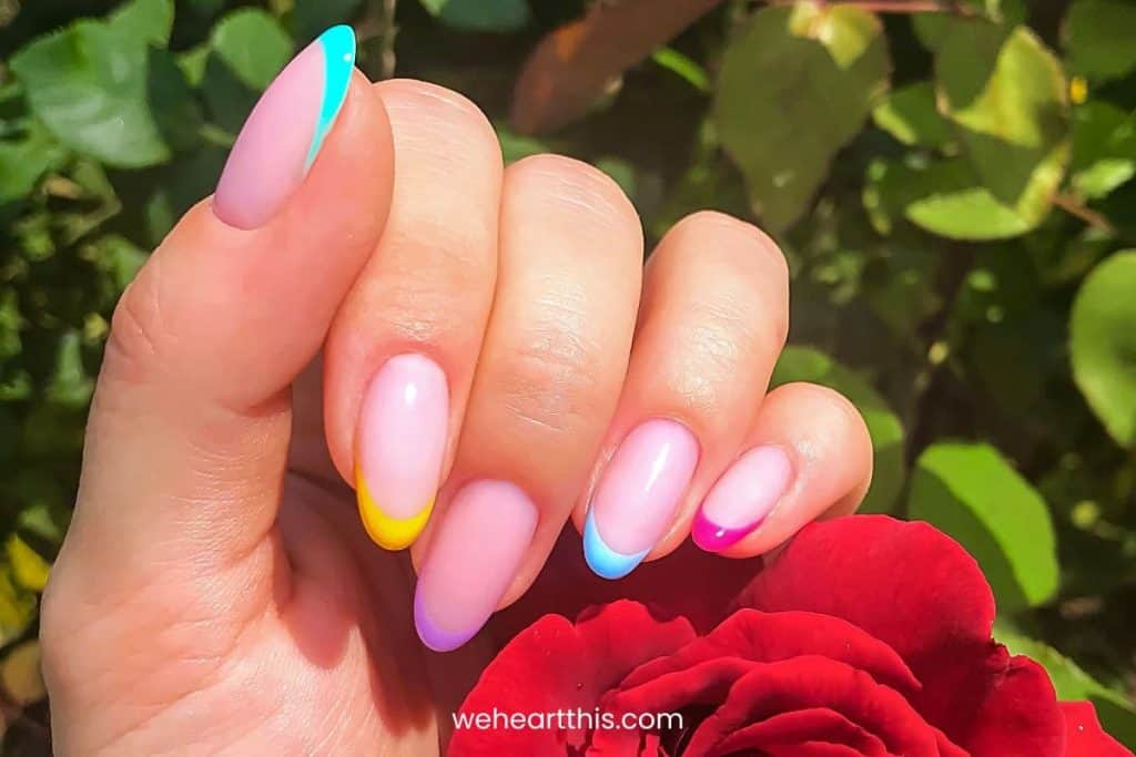 fingernails with pastel colored french tip nails with a red rose and greenery in the background