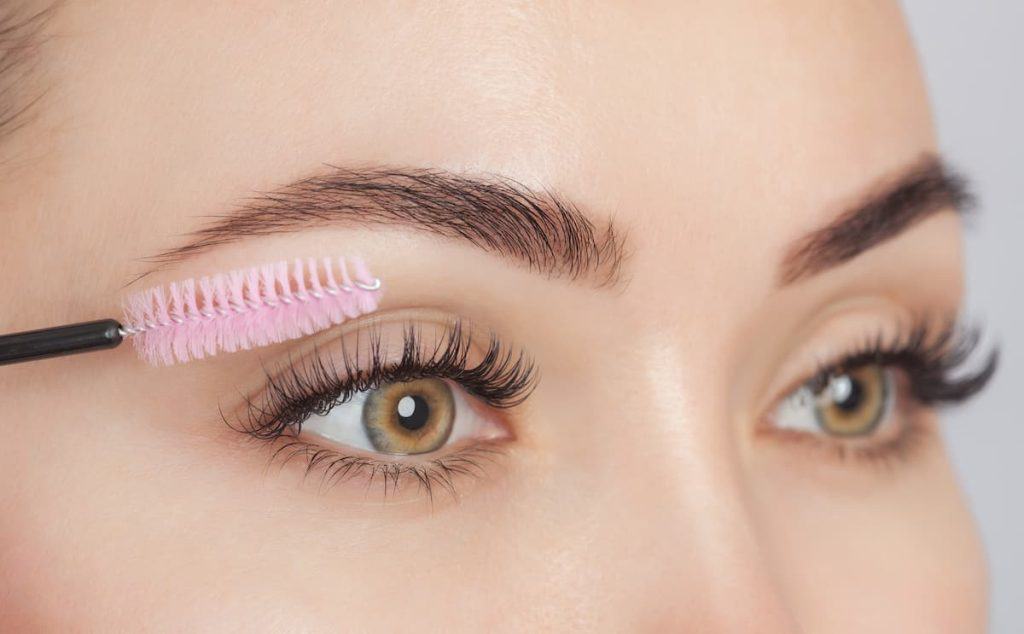 A woman is using a pink spoolie to brush her eyelash extension