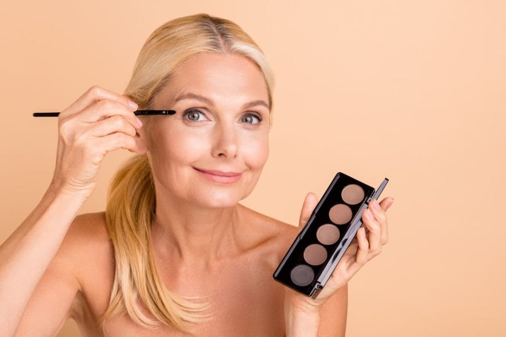 An older woman is holding eyeshadow brush near her eye and eyeshadow palette on other hand while looking at the camera