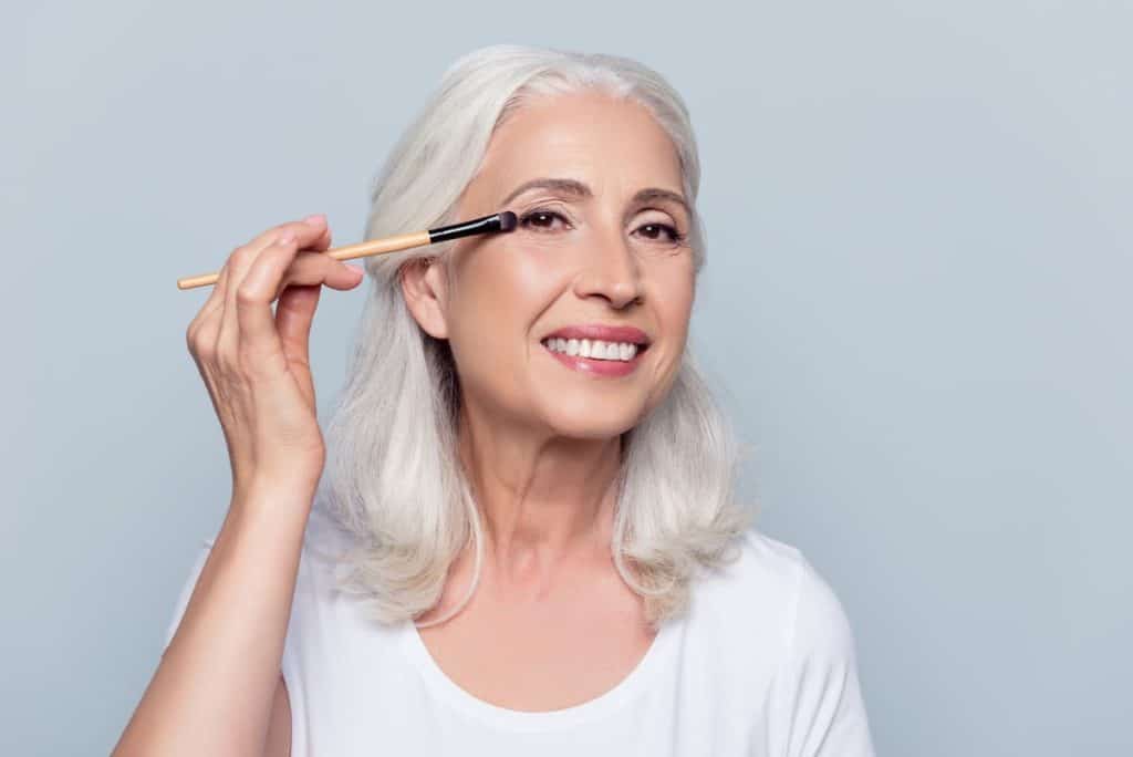 A senior woman is putting eyeshadow on her eyes while smiling and looking at the camera