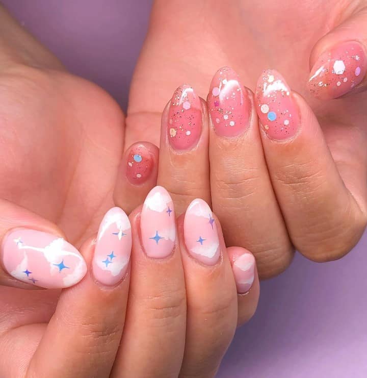 A closeup of a woman's hands with light pink nail polish on one hand and dark pink nail polish on the other that has elements like clouds, stars, and glittery sequins 