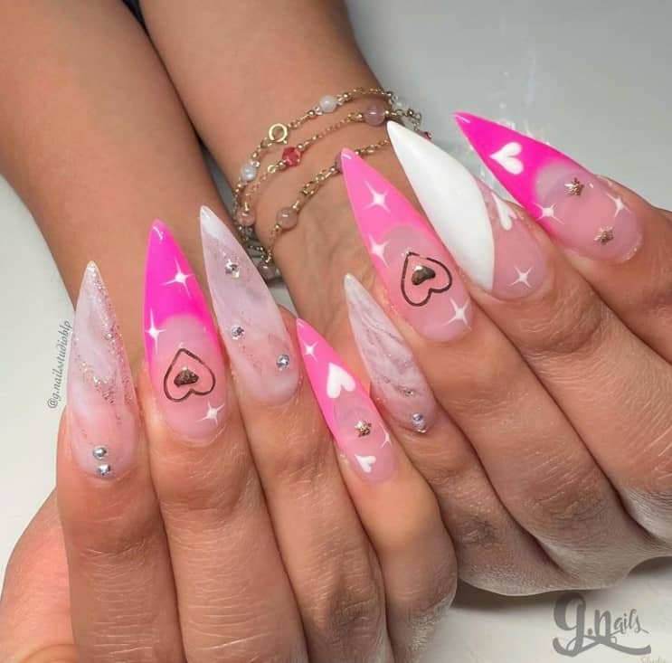 A closeup of a woman's hands with a stiletto acrylics nails that has white hearts, pink French tips, silver rhinestones, white stars, and smoky marble designs