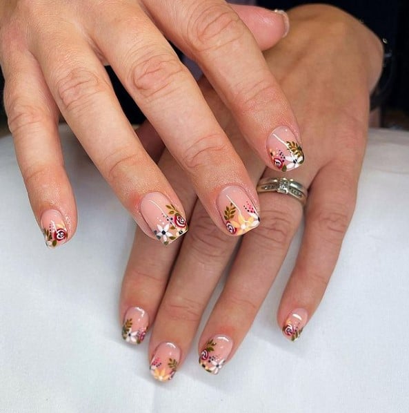 A closeup of a woman's hands with nude nail polish base that has floral French tips with white and red blooms and green foliage