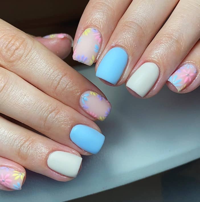 A closeup of a woman's hands with a combination of matte white and blue nail polish that has colorful blooms on select nails