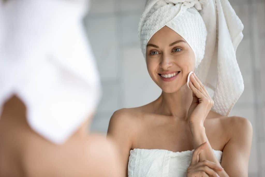 A woman wearing a towel is looking at herself in the mirror while applying facial toner.