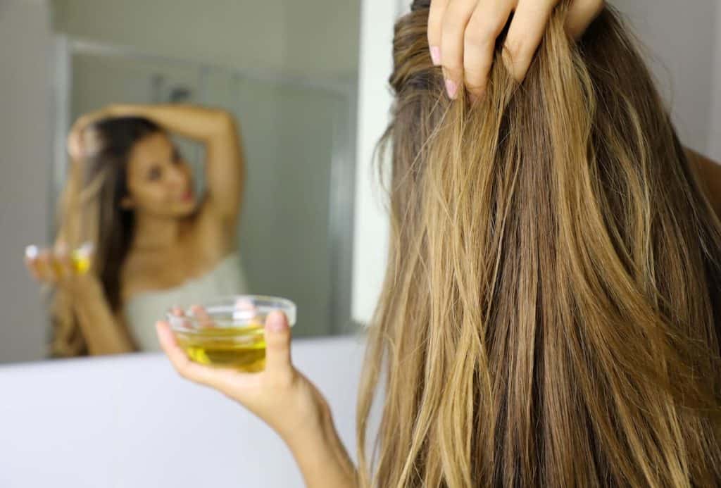 A woman is holding her hair and a bowl of oil on the other hand in front of the mirror