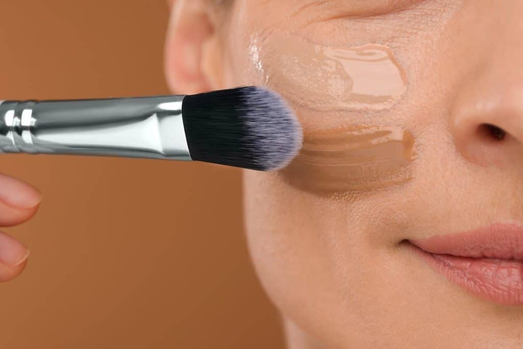A woman is using a brush to apply water-based foundation to her face.