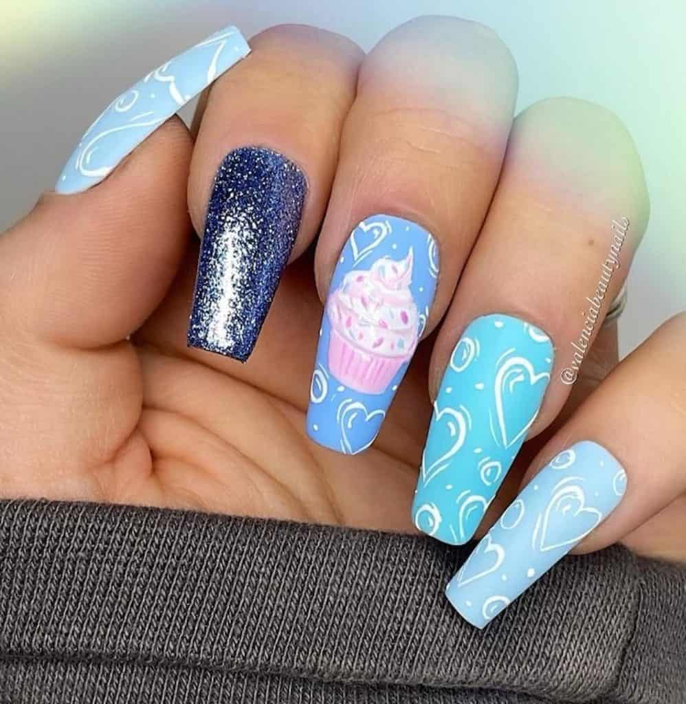 A closeup of a woman's hand with four different shades of blue nail polish that has hand-drawn white hearts, shimmering silver glitter, and pink cupcake nail design