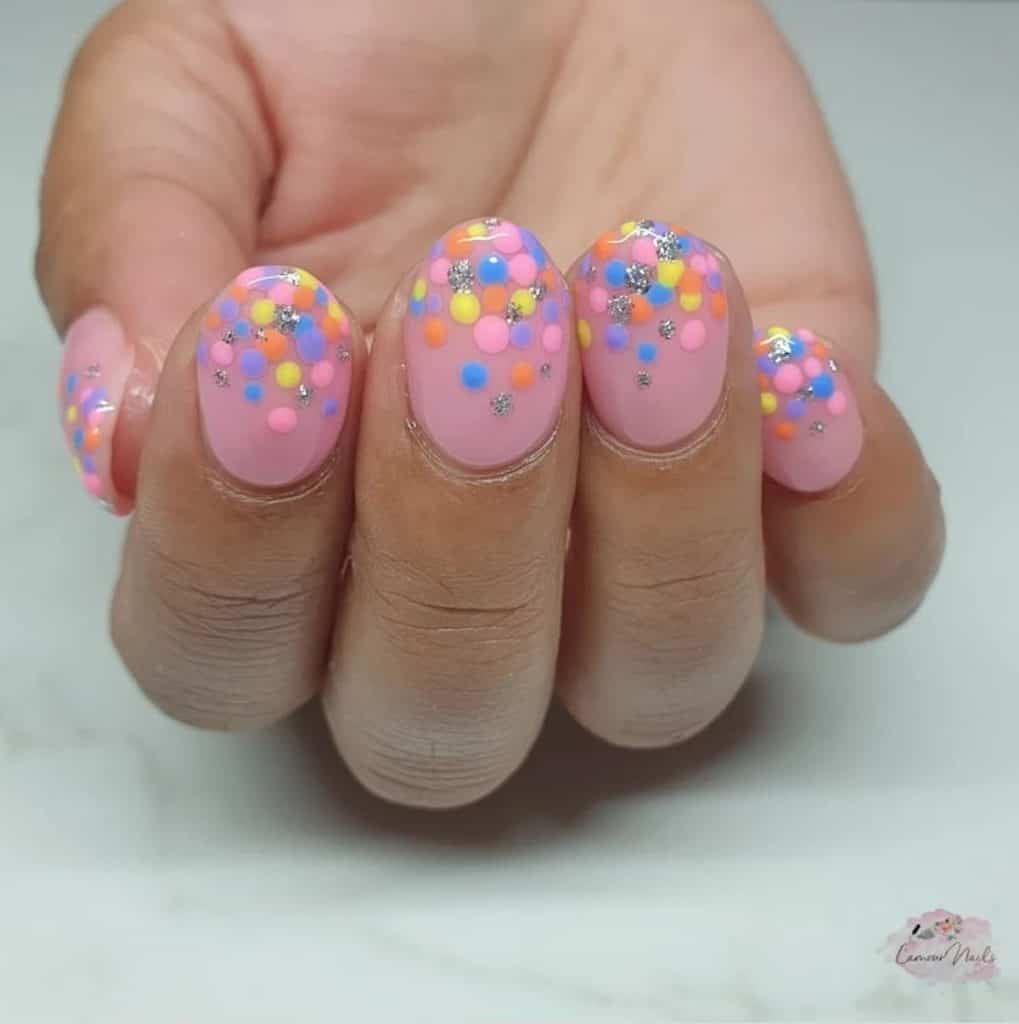 A closeup of a woman's hand with pink nail polish base that has bright and colorful dots and silver glitter accents