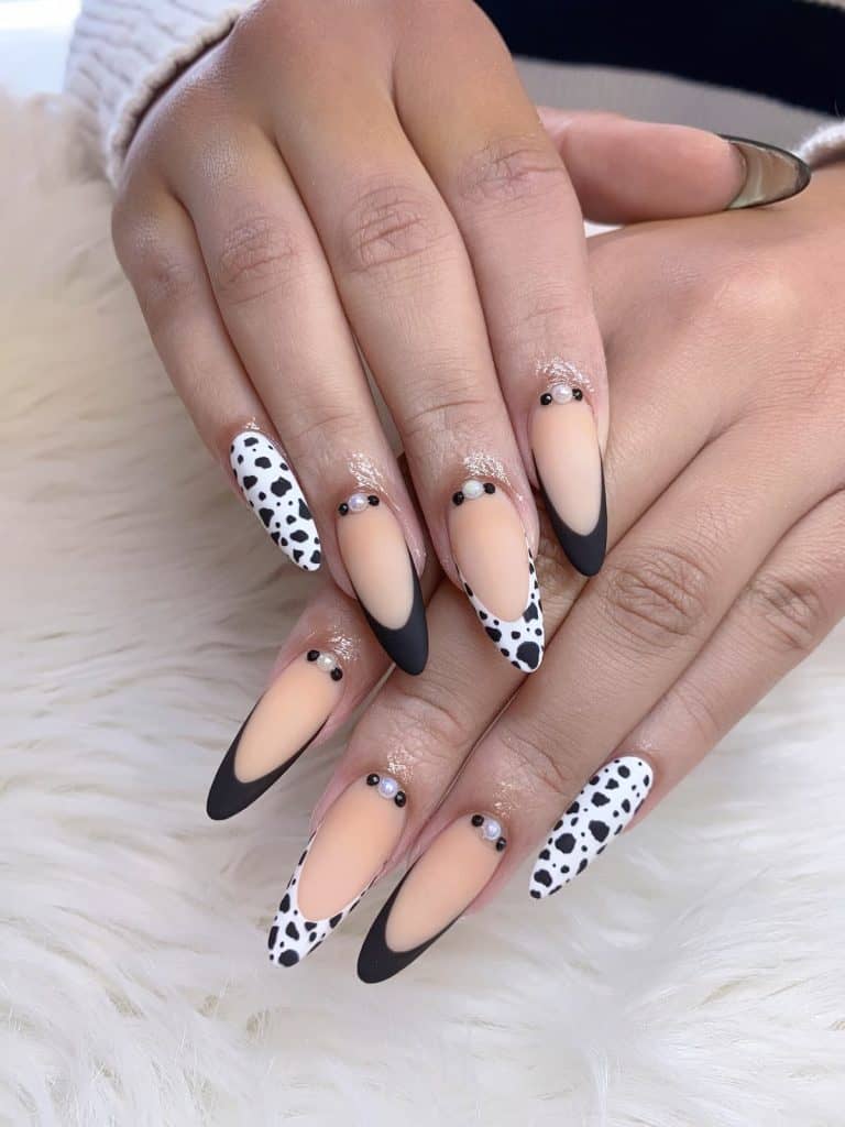 A woman's hands with a combination of beautiful nude, white and black nail polish that has cow print accents, black tips and added jewels on the nails 
