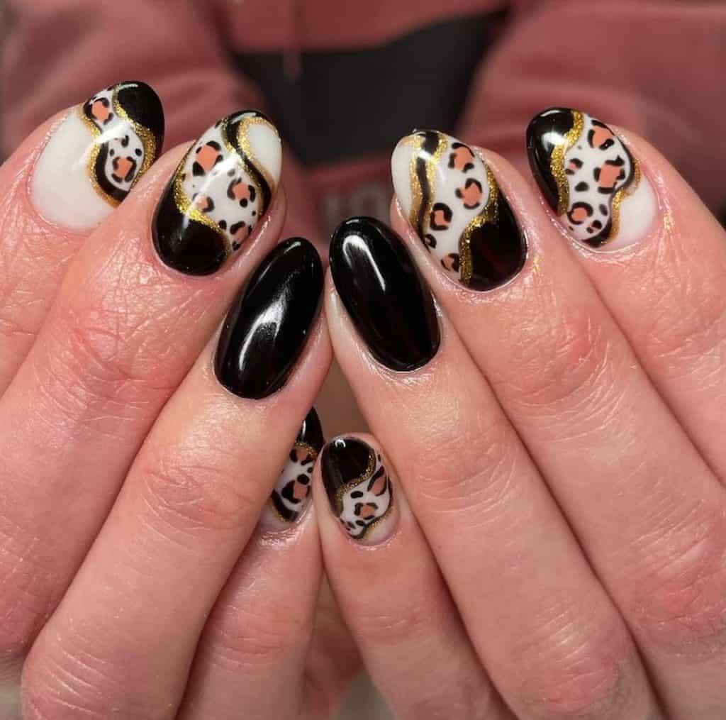 A closeup of a woman's hand with a combination of black and white nail polish that has a leopard nail print design, glittery gold swirls and glossy finish