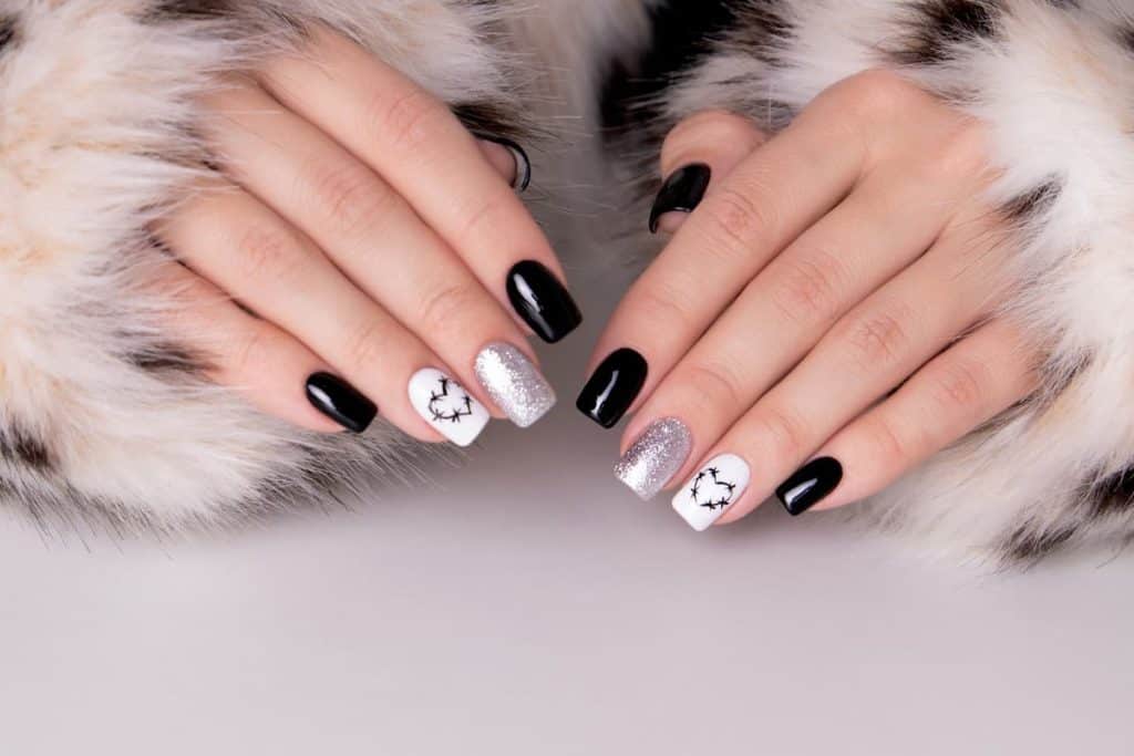 A woman's hands with a combination of beautiful white, black and silver nail polish that has heart with thorns nail designs and silver glitter nails