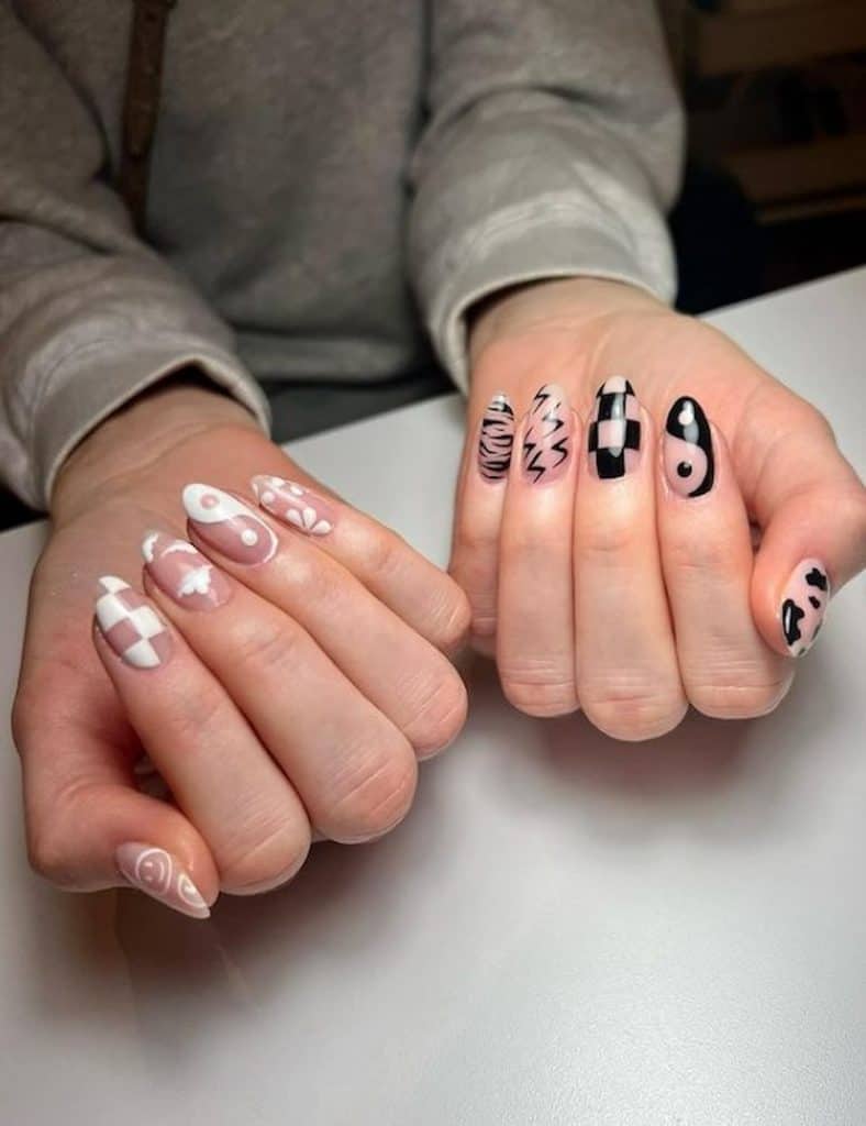 A woman's hands with a combination of nude, black and white nail polish that has yin  yang symbols, floral, clouds, lightning bolts  and checkered pattern nail designs 