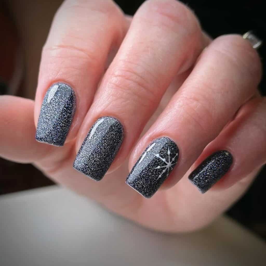 A closeup of a woman's hand with a black nail polish base that has silver glitter and snowflake accent on select nails