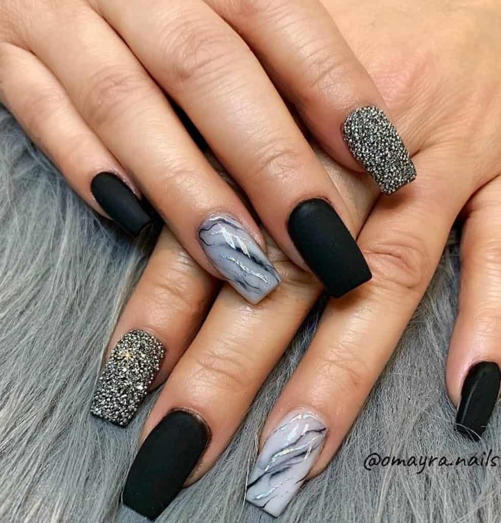 A closeup of a woman's hands with matte black nail polish that has white glitter and glossy marble acrylic nails
