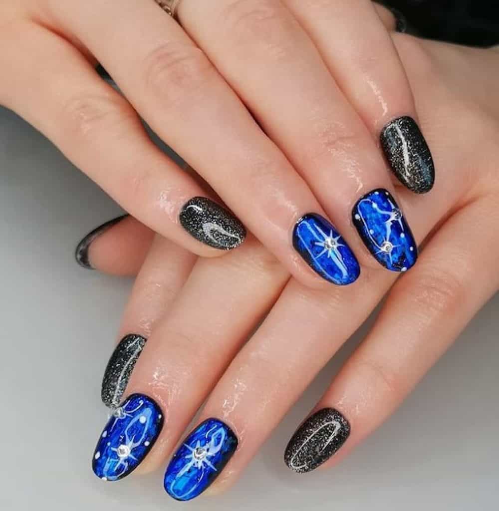 A woman's hands with glittery black and sapphire nail polish that has white details and rhinestones 