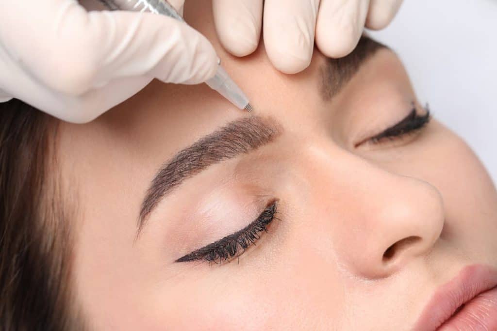 Close up photo of a young woman with her eyes closed during procedure of permanent eyebrow makeup