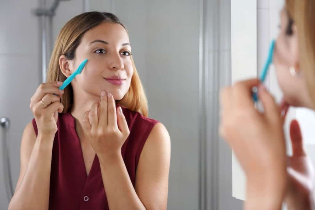 A woman using razor near her face while looking at herself in the mirror on the bathroom