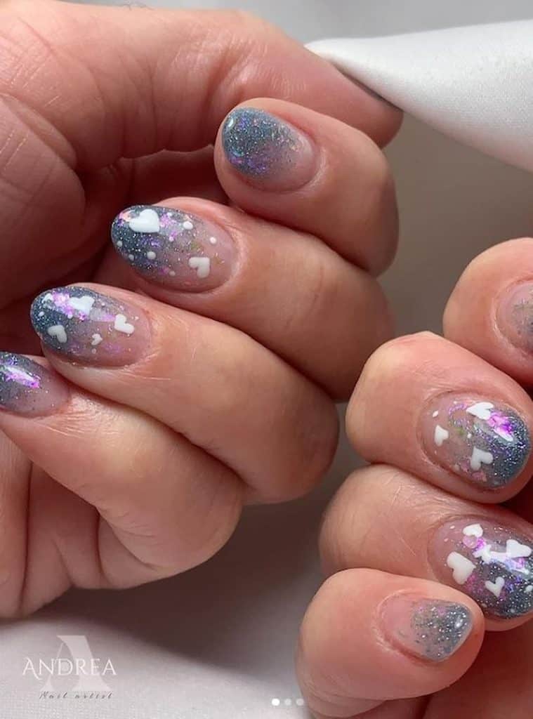 A closeup of a woman's nails with a combination of blue-and-bare ombré nail polish that has glitters and white heart nail designs