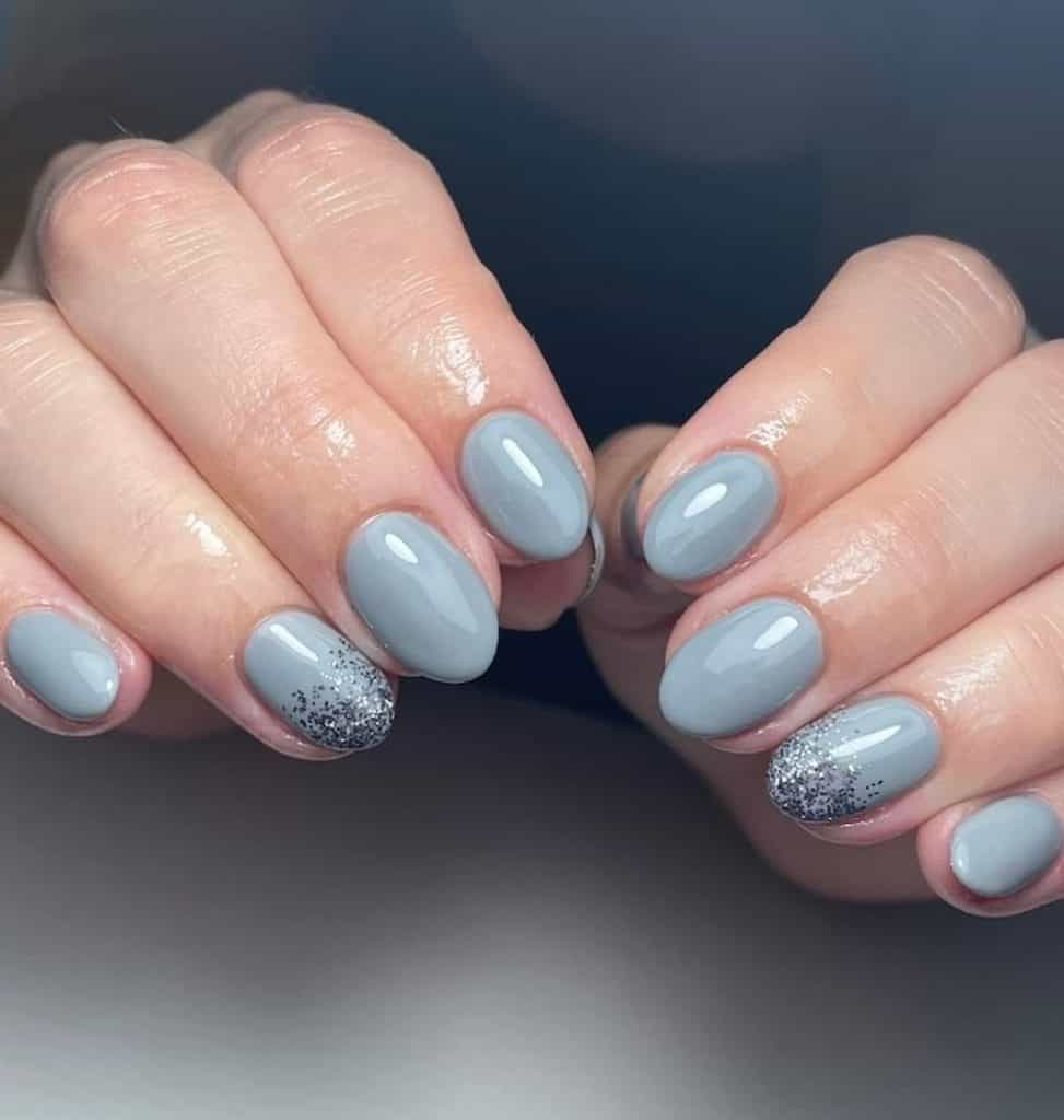 A closeup of a woman's hands with slate blue nail polish base that has an ombré of silver glitter nail designs on select nails