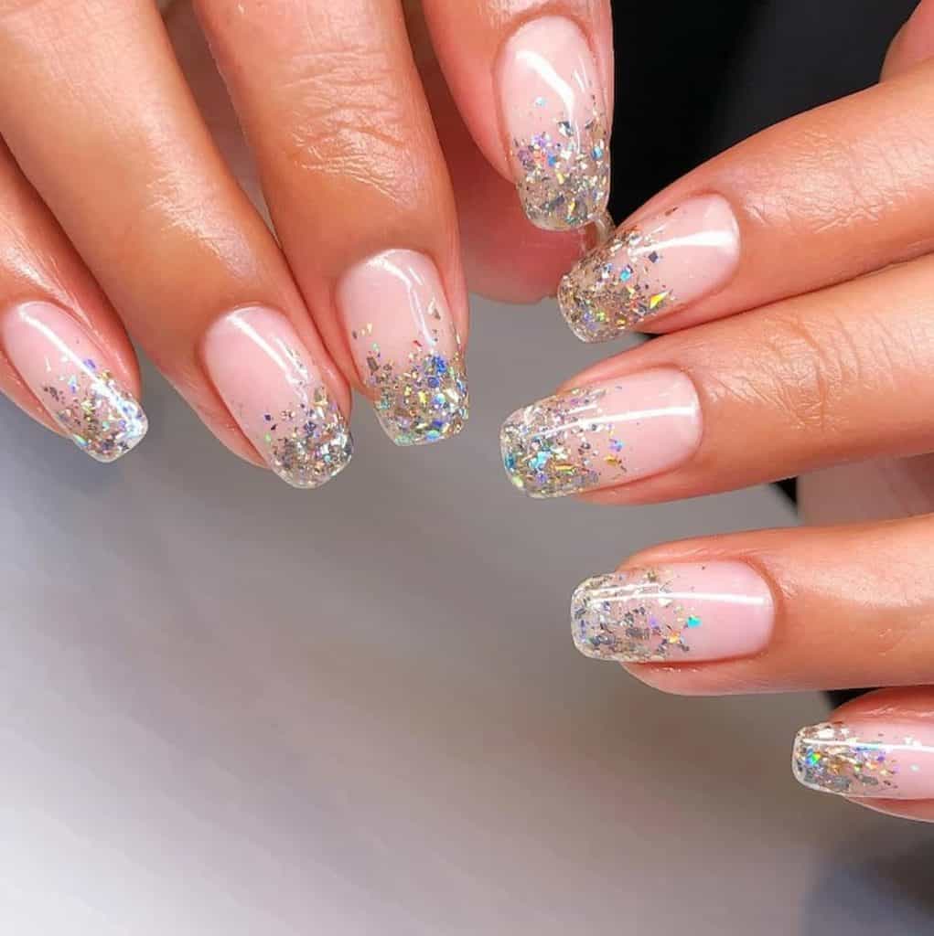 A closeup of a woman's hands with a glossy nude nail polish that has holographic glitters nail designs on the tips of nails