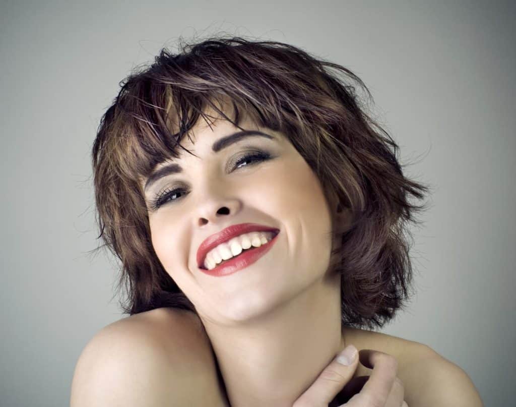 A closeup of a brunette short haired woman, wearing a makeup, smiling. Isolated on a gray background