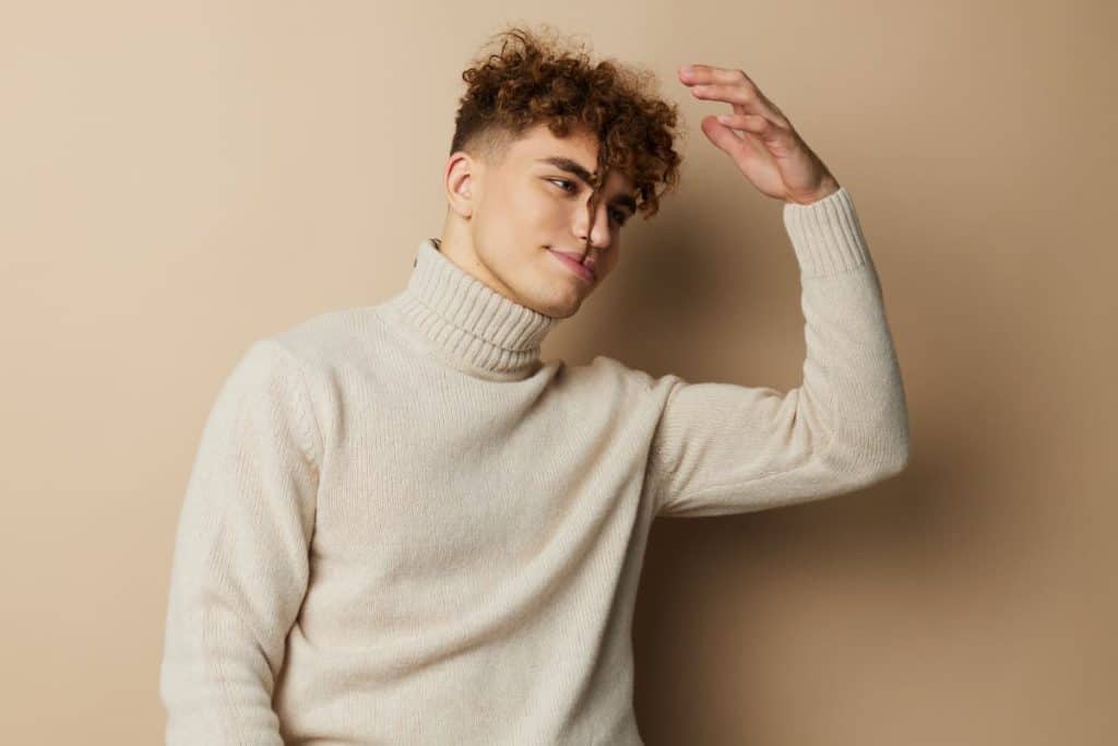 A man with curly top of his hair wearing a light-colored turtle neck sweater isolated on a brown background
