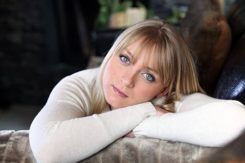 A blonde woman with wispy bangs and blue eyes, sitting on a couch