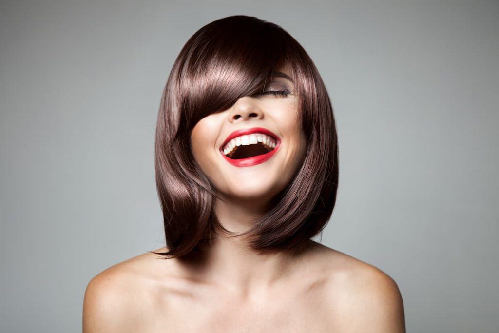 A woman with a brown bob and red lipstick is laughing with her mouth open on a gray background.