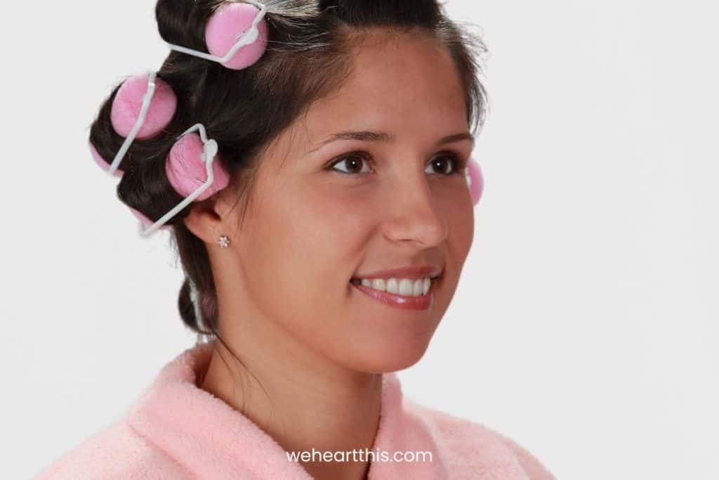 A woman wearing pink robe and has foam hair rollers on her hair, smiling while looking at the side. Isolated on a white background