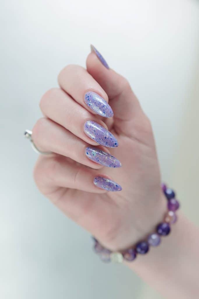 A woman's hand wearing a bracelet with a long, gleaming almond lavender nails and shiny blue flakes 