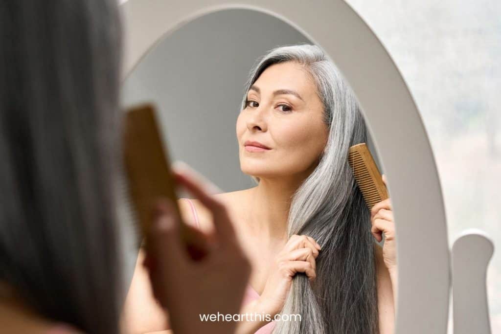 A mature woman with gray hair using a comb to her hair while looking at herself in the mirror