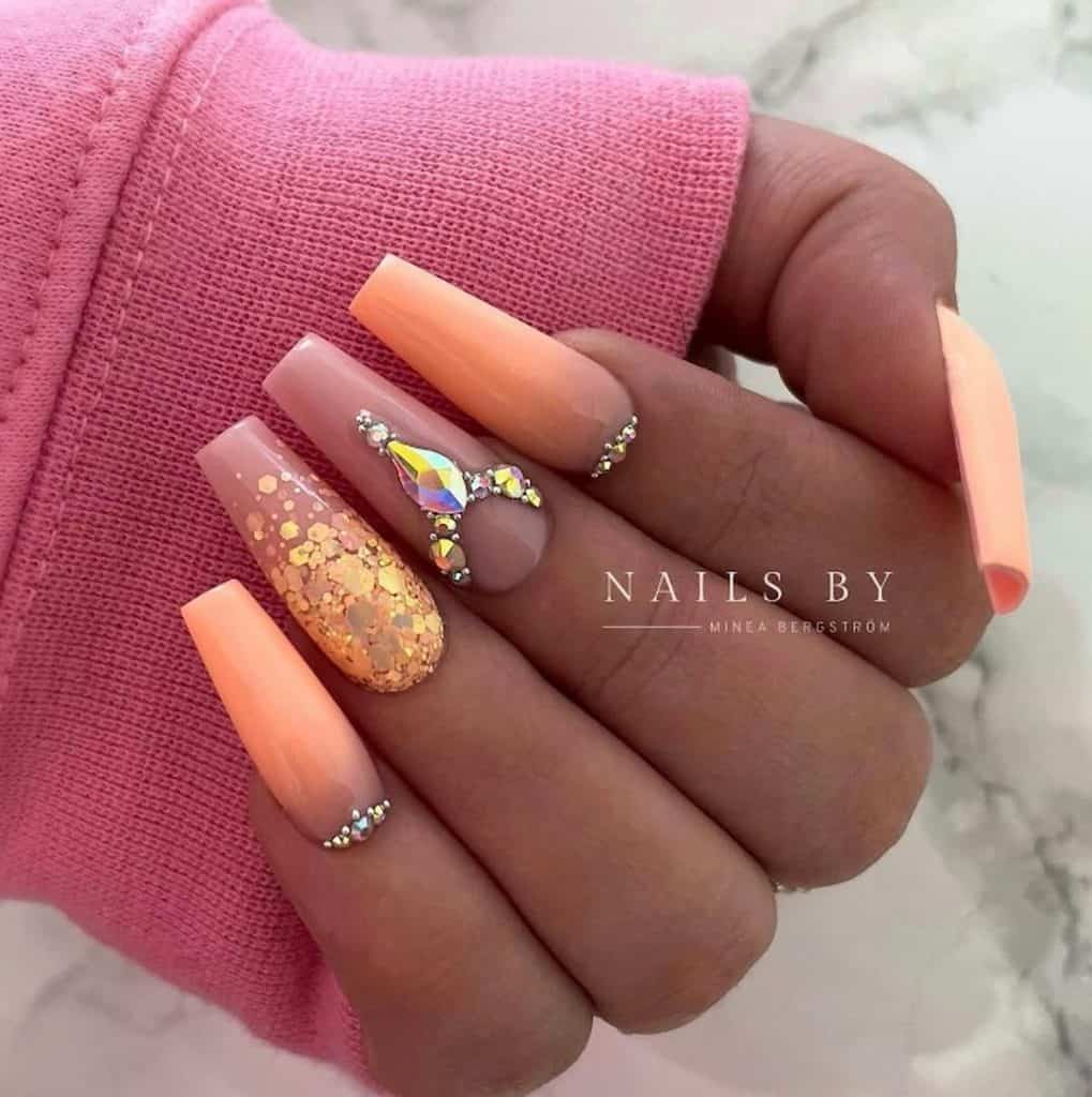 A closeup of a woman's hand with a nude pink and warmer peach nail polish that has peach sequins and gem accents 