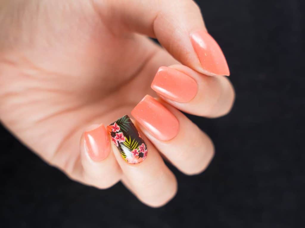 A closeup of a woman's hands with peach nail polish base that has tropical-themed print against a black background on one fingernail