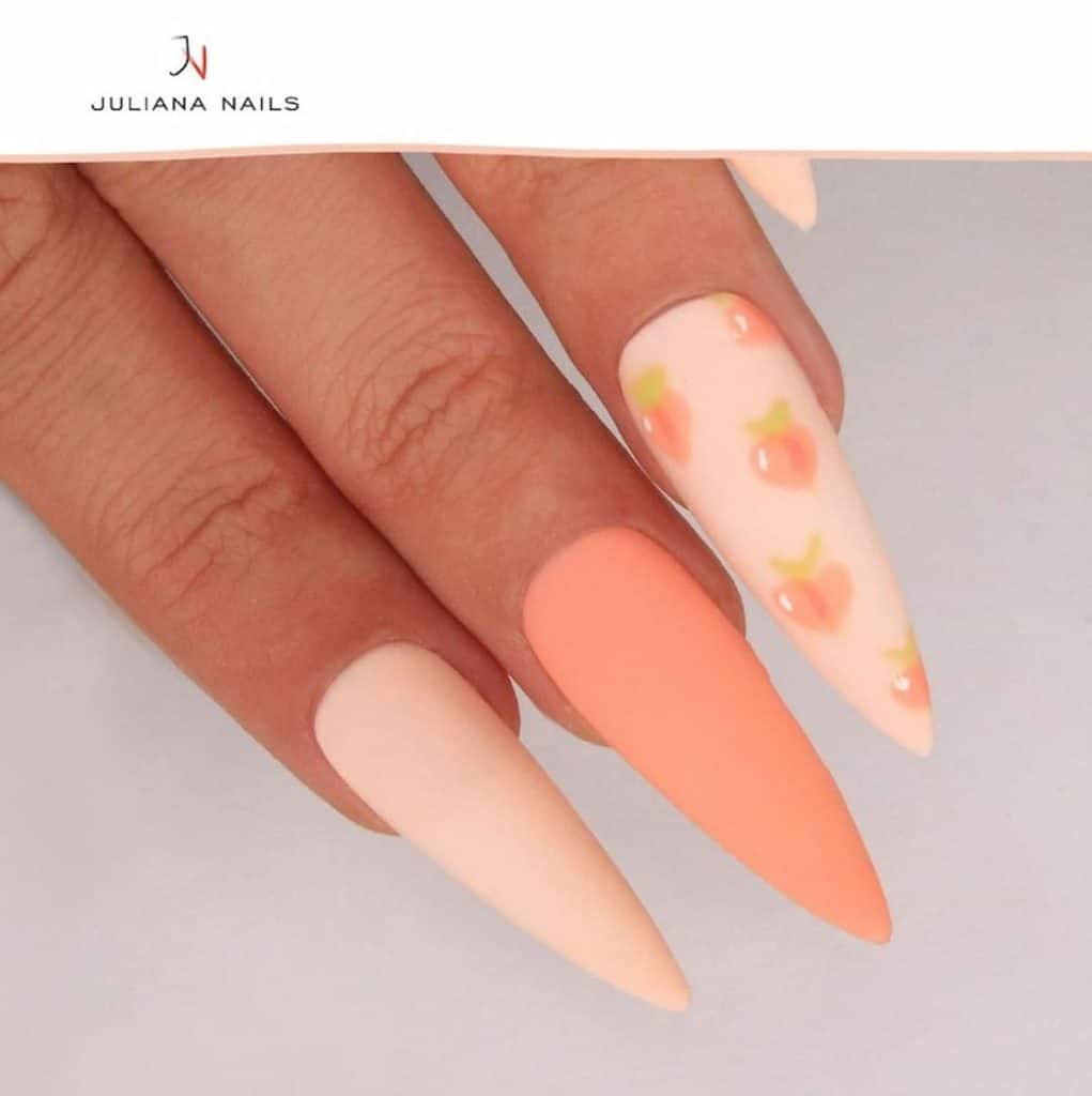 A closeup of a woman's hands with matte peach nail polish and long stiletto nails that has peach fruits nail designs on select nails