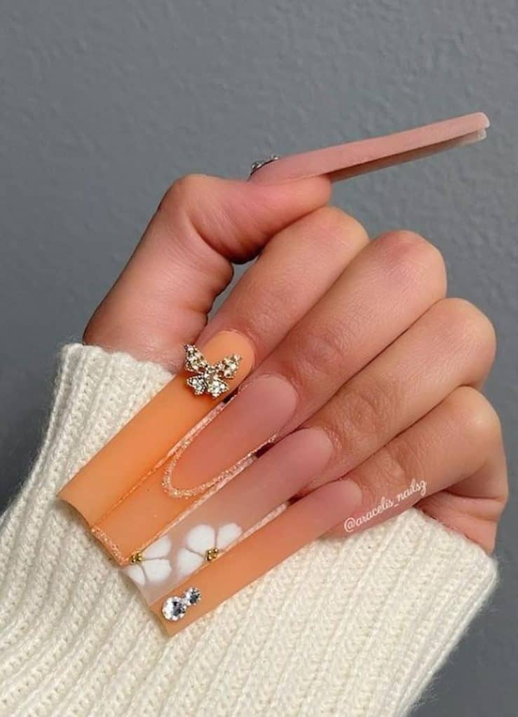 A closeup of a woman's hand with a long square nails in gradient peach that has gems, studs, florals, and glitter-outlined French tips nail designs