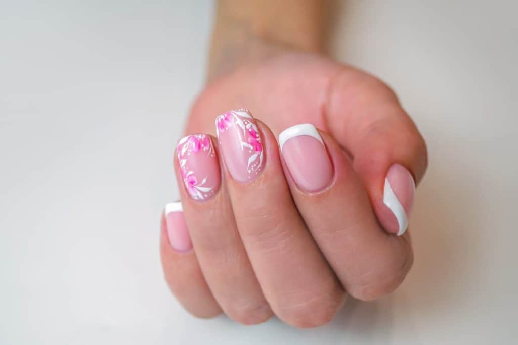A closeup of a woman's hand with a pink nail polish and white tips that has white floral borders nail designs