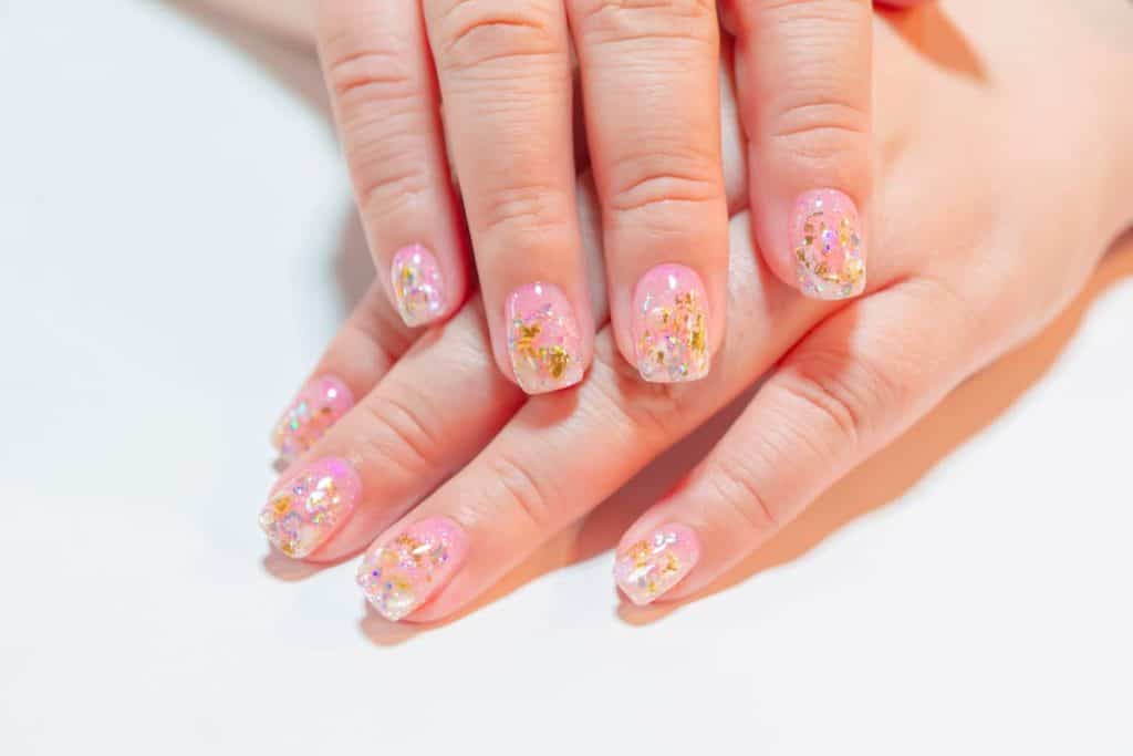 A closeup of a woman's hands with pink nail polish that has gold and silver flakes nail designs