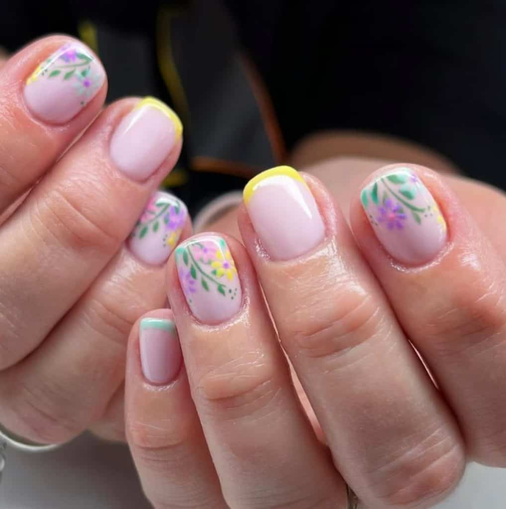 A closeup of a woman's hands with nude nail polish and yellow nail tips on select nails that has multicolored floral and leaves nail designs on select nails