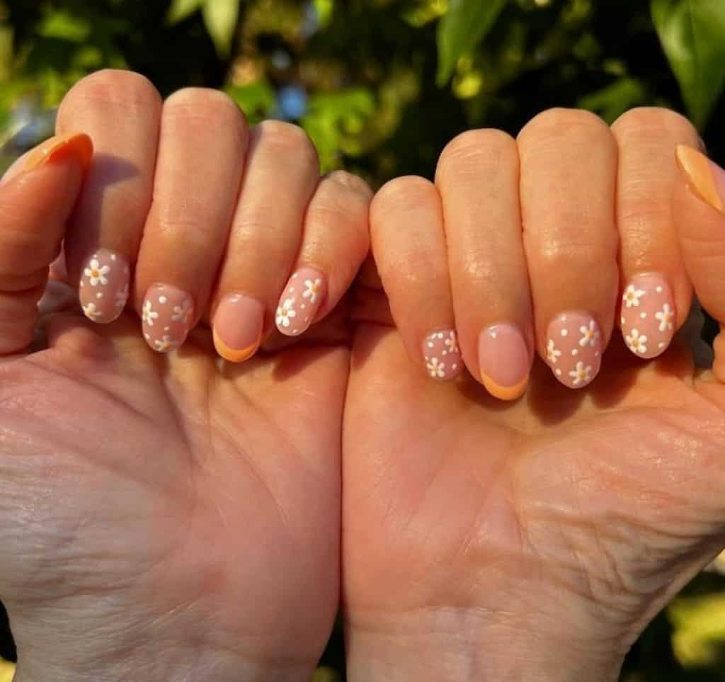 A closeup of a woman's hands with nude nail polish and orange tips on select nails that has flowers nail designs