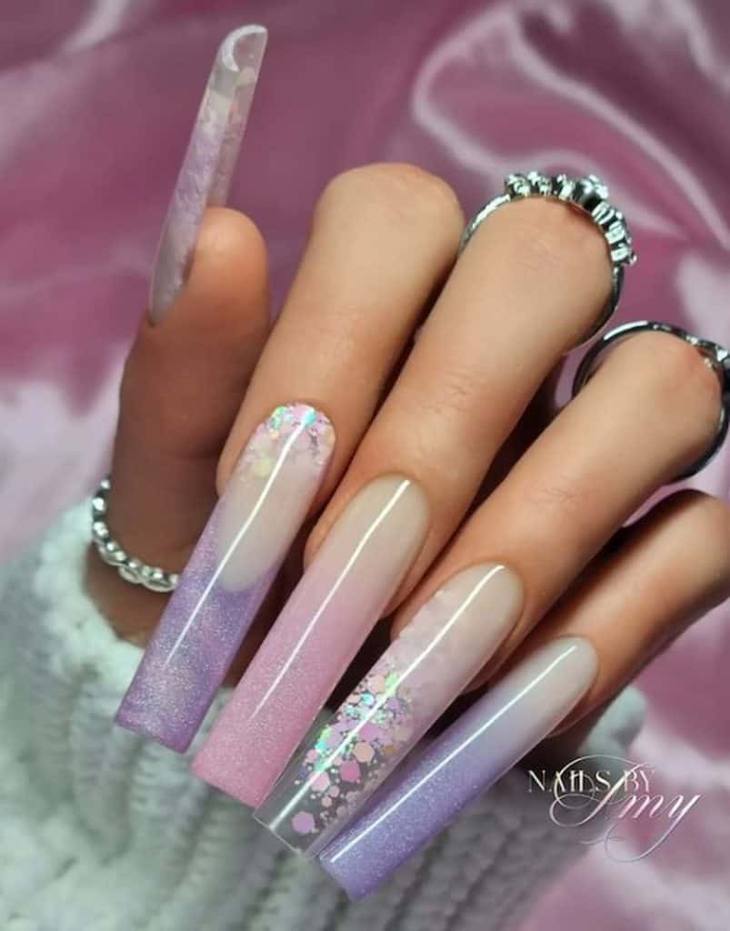 A closeup of a woman's hand with long nails that features a layering of soft pink and lavender glitters and sequins nail designs