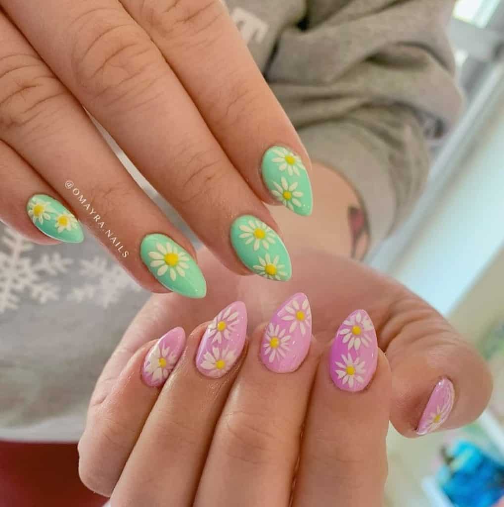 A closeup of a woman's hands withalmond-shaped acrylic nails in green on one hand and pink on the other that has daisies nail designs