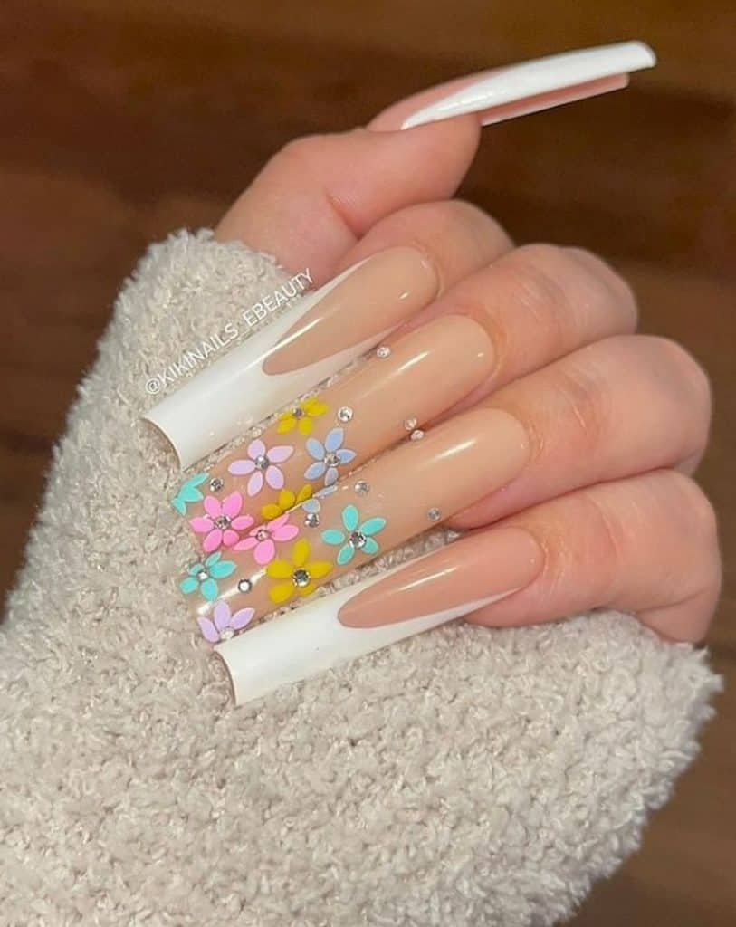 A closeup of a woman's hand with a long classic French manicure, nude nail polish base, and white tips on select nails that has multicolored flowers and glittery studs nail designs