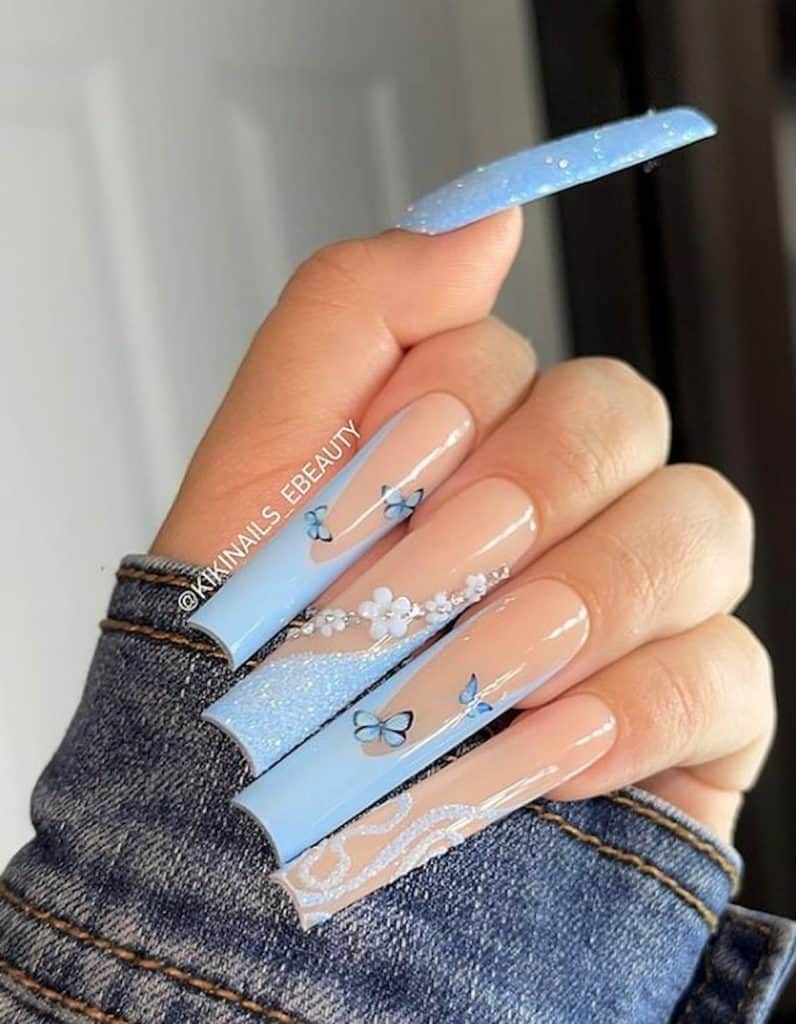 A closeup of a woman's hand with extra long ballerina nails and nude nail polish base that has light blue-colored French tips, blue sugary glitters, blue butterflies and 3D flowers nail designs
