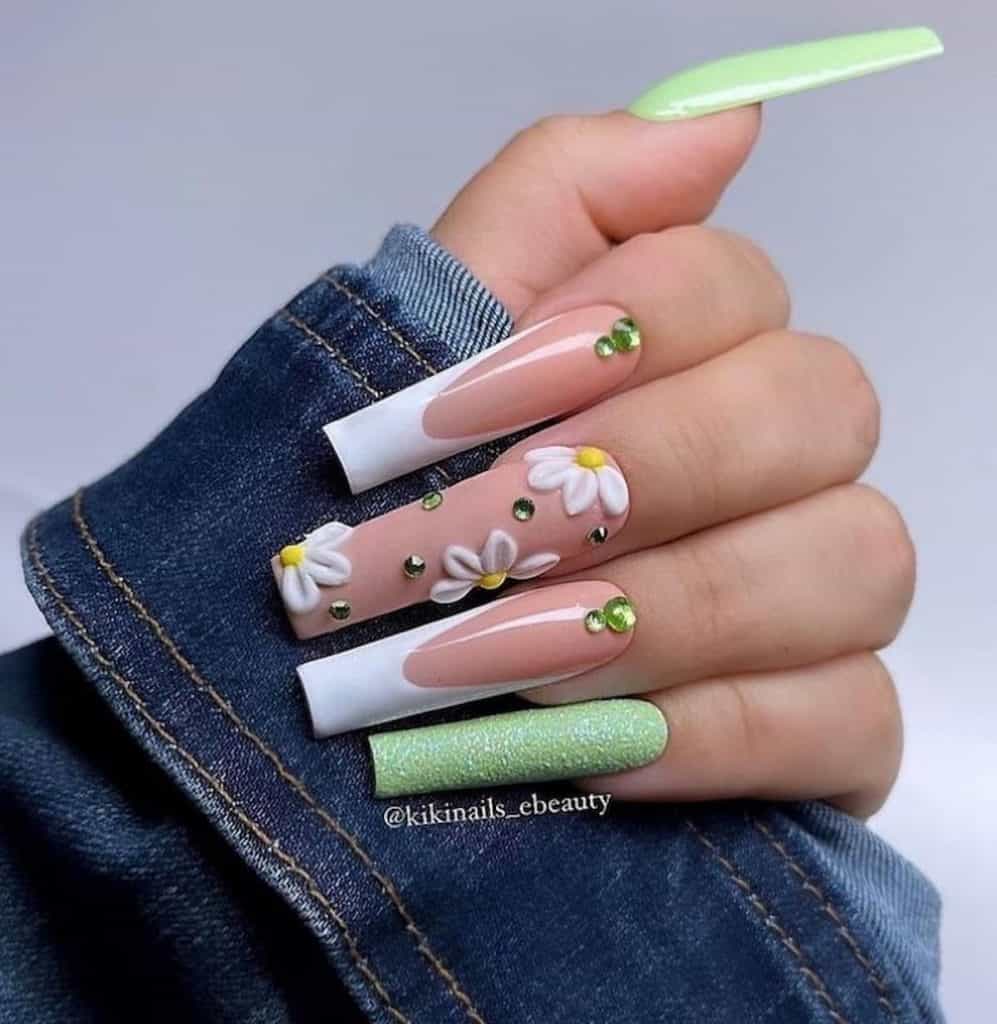 A closeup of a woman's hand with a combination of green and nude nail polish that has white tips on select nails and 3D daisies and green studs nail designs