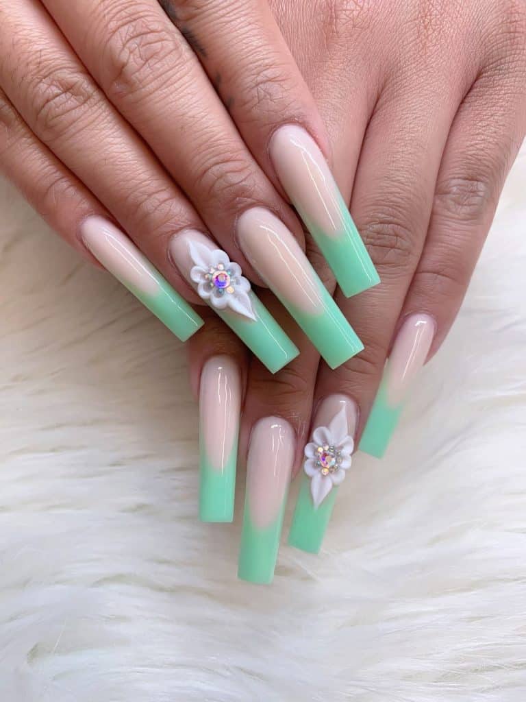 A closeup of a woman's hands with long nails and mint green nail tips that has studded 3D flower in white nail designs on select nails