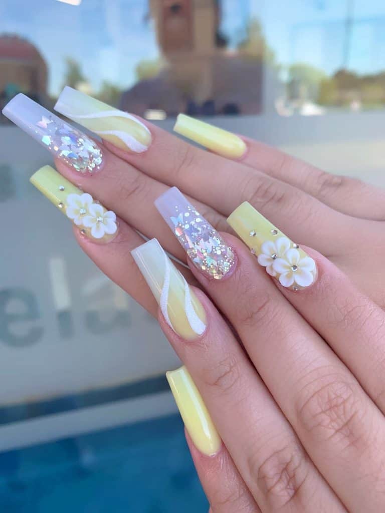 A closeup of a woman's hands with a pastel yellow nail polish that has holographic sequins and butterflies on sheer lavender, white squiggly patterns, 3D florals, and studs nail designs