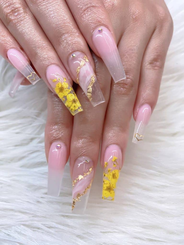 A woman's hands with a combination of pink-and-clear gradient acrylics that has tiny stud, golden and marble streaks, yellow florals, and tiny golden hearts nail designs